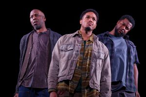 Three men stand on stage with serious expressions all looking in different directions