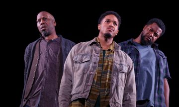 Three men stand on stage with serious expressions all looking in different directions