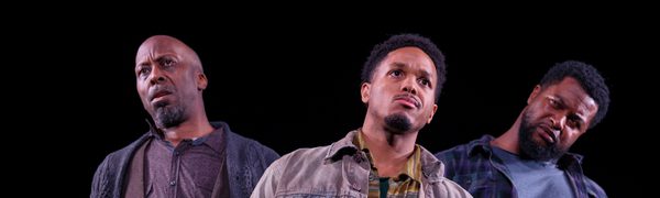 Three men stand on stage with serious expressions all looking in different directions
