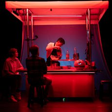 A person works at a food stand illuminated by red light as two people sit by the counter on stools