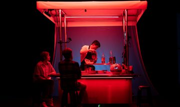 A person works at a food stand illuminated by red light as two people sit by the counter on stools