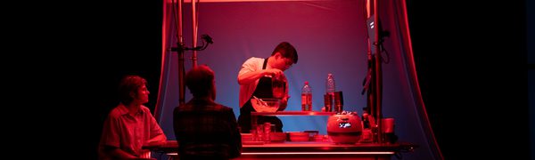 A person works at a food stand illuminated by red light as two people sit by the counter on stools