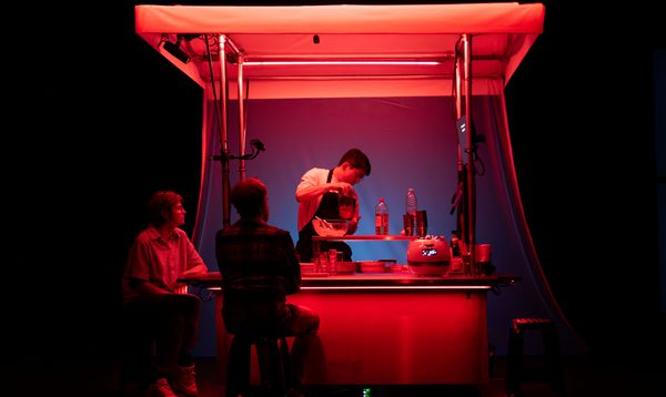 A person works at a food stand illuminated by red light as two people sit by the counter on stools
