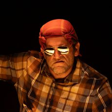 A grumpy looking man wearing sunglasses and a bandana holding up a wanted poster with a drawing that looks like him