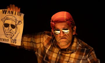 A grumpy looking man wearing sunglasses and a bandana holding up a wanted poster with a drawing that looks like him