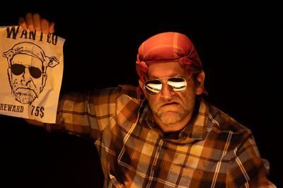 A grumpy looking man wearing sunglasses and a bandana holding up a wanted poster with a drawing that looks like him