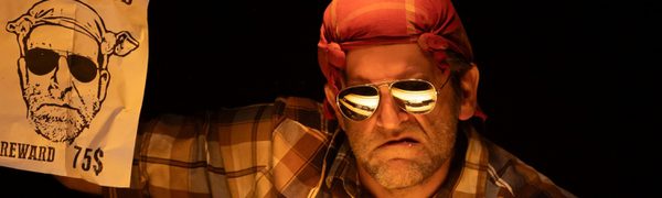 A grumpy looking man wearing sunglasses and a bandana holding up a wanted poster with a drawing that looks like him