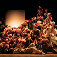 A large ensemble of dancers move as one while tightly packed together and holding white and red flowers