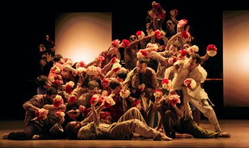 A large ensemble of dancers move as one while tightly packed together and holding white and red flowers