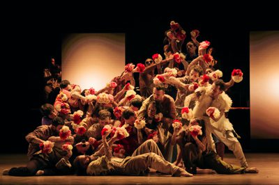 A large ensemble of dancers move as one while tightly packed together and holding white and red flowers
