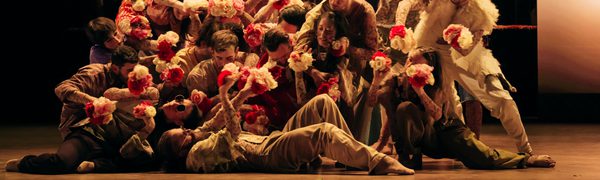 A large ensemble of dancers move as one while tightly packed together and holding white and red flowers
