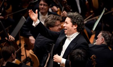 A conductor standing with one arm raised and a smile surrounded by orchestra members