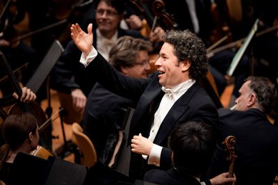 A conductor standing with one arm raised and a smile surrounded by orchestra members