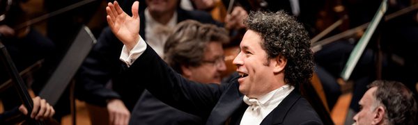 A conductor standing with one arm raised and a smile surrounded by orchestra members