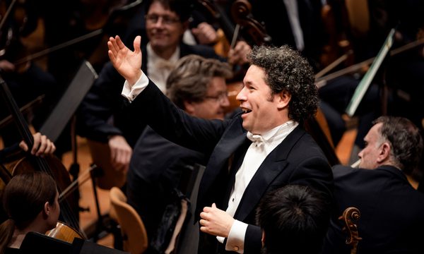 A conductor standing with one arm raised and a smile surrounded by orchestra members