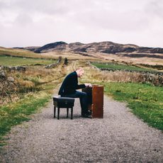 A man sitting at a keyboard instrument in the middle of a country path surrounded by fields and hills