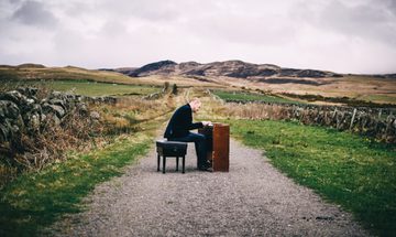 A man sitting at a keyboard instrument in the middle of a country path surrounded by fields and hills