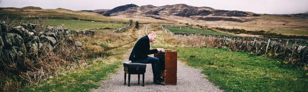 A man sitting at a keyboard instrument in the middle of a country path surrounded by fields and hills