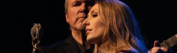 a women sings with a man playing guitar behind her