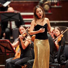 A woman sings on stage wearing a black top and a gold sparkly skirt while an orchestra plays behind her