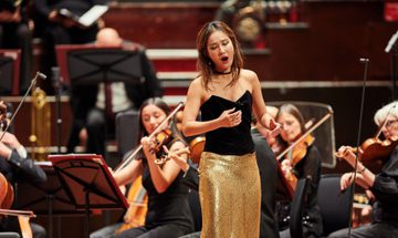 A woman sings on stage wearing a black top and a gold sparkly skirt while an orchestra plays behind her