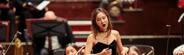 A woman sings on stage wearing a black top and a gold sparkly skirt while an orchestra plays behind her
