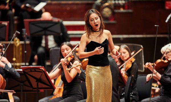 A woman sings on stage wearing a black top and a gold sparkly skirt while an orchestra plays behind her