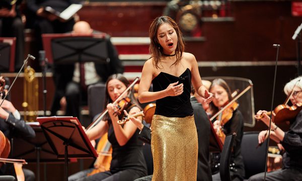 A woman sings on stage wearing a black top and a gold sparkly skirt while an orchestra plays behind her
