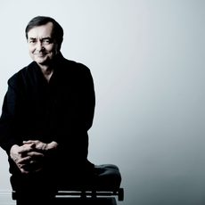 A man dressed in black sits in front of a white wall smiling at the camera