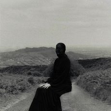 A black and white image of a person covered head to toe in black clothes is positioned in a seated position in the middle of a rural road