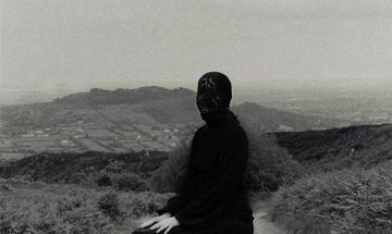 A black and white image of a person covered head to toe in black clothes is positioned in a seated position in the middle of a rural road