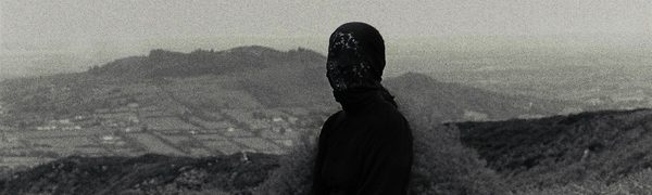 A black and white image of a person covered head to toe in black clothes is positioned in a seated position in the middle of a rural road