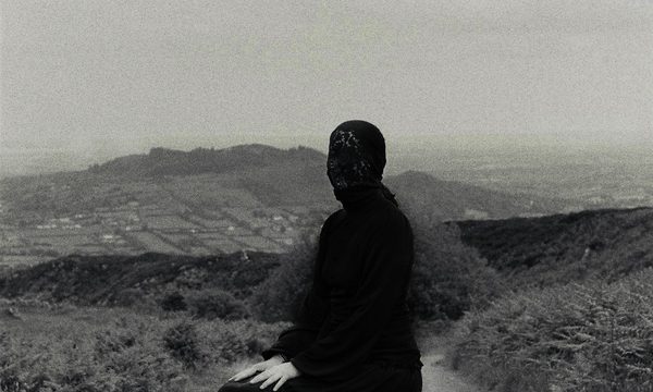 A black and white image of a person covered head to toe in black clothes is positioned in a seated position in the middle of a rural road