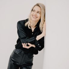 A woman dressed in black leans against a white wall while smiling into the distance