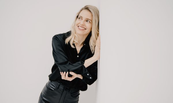 A woman dressed in black leans against a white wall while smiling into the distance