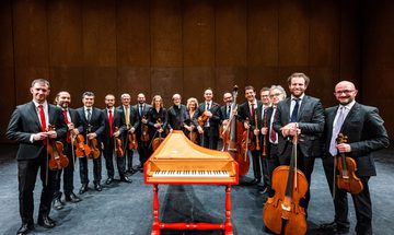 The Venice Baroque orchestra holding their instruments while standing around a keyboard instrument