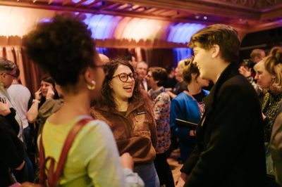 Two women stand chatting in The Hub Main Hall amidst a crowd of people, both laughing and smiling.