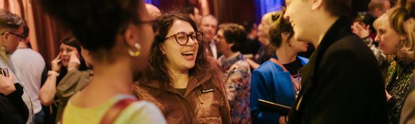 Two women stand chatting in The Hub Main Hall amidst a crowd of people, both laughing and smiling.