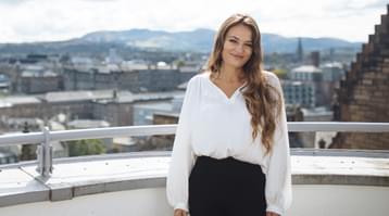 Nicola Benedetti stands on a balcony smiling at the camera with a view of Edinburgh behind her
