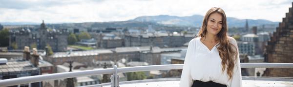 Nicola Benedetti stands on a balcony smiling at the camera with a view of Edinburgh behind her