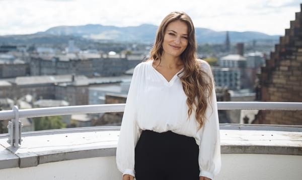 Nicola Benedetti stands on a balcony smiling at the camera with a view of Edinburgh behind her