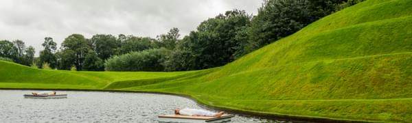 Person lying on a planks of wood in water surrounded by green sculptured landscape of Jupiter Artland.