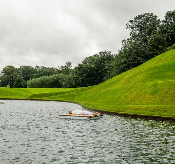 Person lying on a planks of wood in water surrounded by green sculptured landscape of Jupiter Artland.