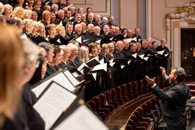 A chorus stand on wooden risers dressed all in black, with a conductor standing in front of them with his arms up