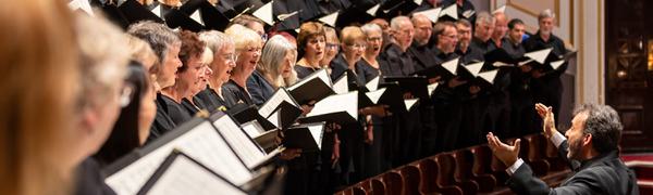 A chorus stand on wooden risers dressed all in black, with a conductor standing in front of them with his arms up