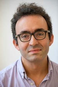 A white man with short dark curly hair and chunky dark-rimmed glasses looks into the camera with a slight smile