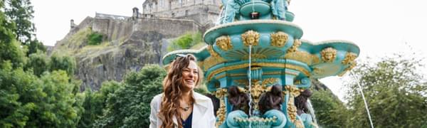 Nicola Benedetti stands smiling in front of a fountain in Princes Street Gardens with Edinburgh Castle visible in the background