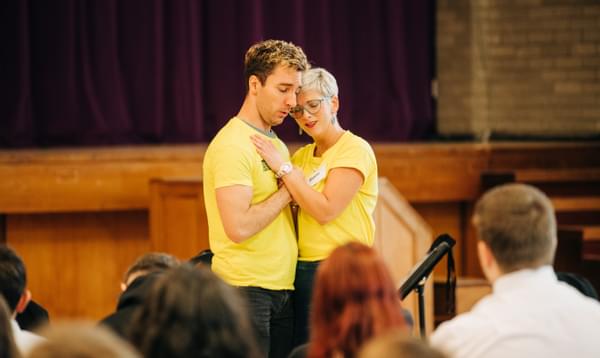 A man and a woman wearing yellow t-shirts embrace each other looking sad