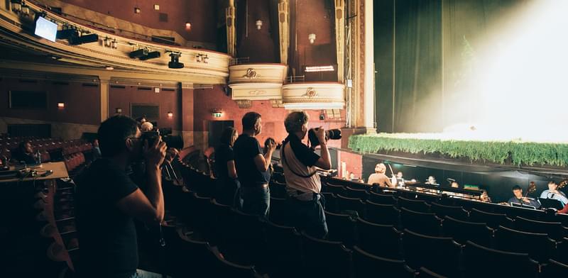 A group of photographers gather together within a theatre auditorium capturing the a brightly lit stage.