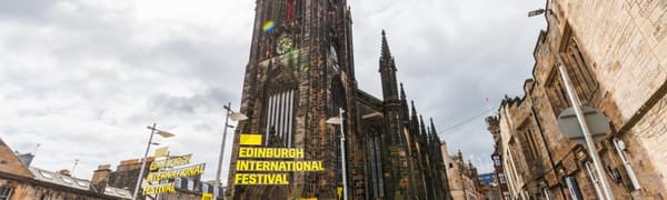 The Hub is surrounded by crowds of people, with a yellow sign reading 'Edinburgh International Festival' floating in front of the spire